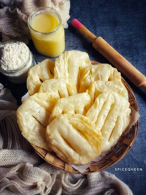 A basket of golden-brown, layered fried dough sticks or fritters, styled with a glass of yellow oil, a jar of white flour, and a wooden rolling pin on a dark blue textured background.