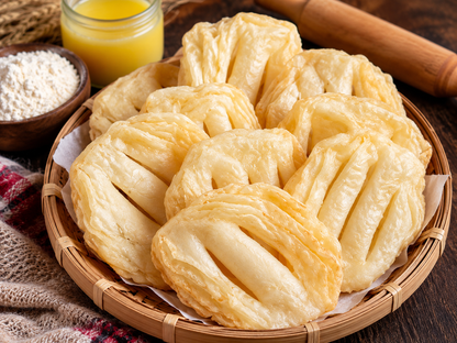 Golden-brown fried pastry cakes arranged in a round bamboo tray, with a small bowl of white flour, a glass of yellow liquid, and a wooden rolling pin on a dark wooden table.