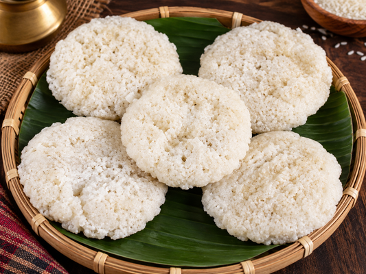 Five round white rice cakes with a porous, crispy texture arranged on a green banana leaf inside a woven bamboo tray, set on a dark wooden table.