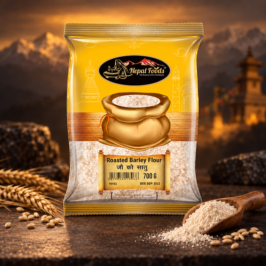 A 700g bag of Roasted Barley Flour stands on a rustic wooden surface with scattered grains and a wooden scoop. The background features a warm, blurred mountain landscape with golden hues.