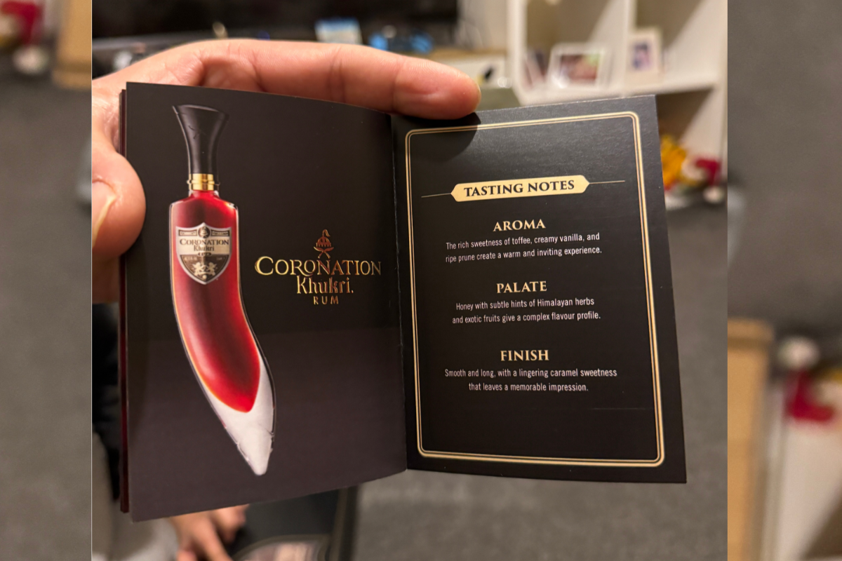 A hand holds an open booklet for the Coronation Khukuri Rum bottle, showing a printed bottle illustration on the left page and tasting notes on the right page.