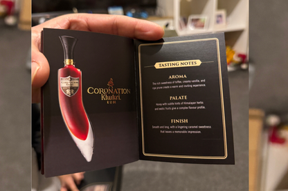 A hand holds an open booklet for the Coronation Khukuri Rum bottle, showing a printed bottle illustration on the left page and tasting notes on the right page.