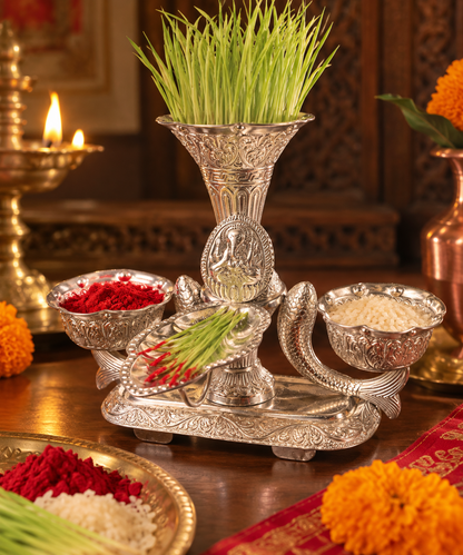 An ornate silver-toned decorative pedestal with carved details, holding green grass, red powder, white grains, and a small plate of green onions, set on a wooden table with warm lit candles and flowers in the background.