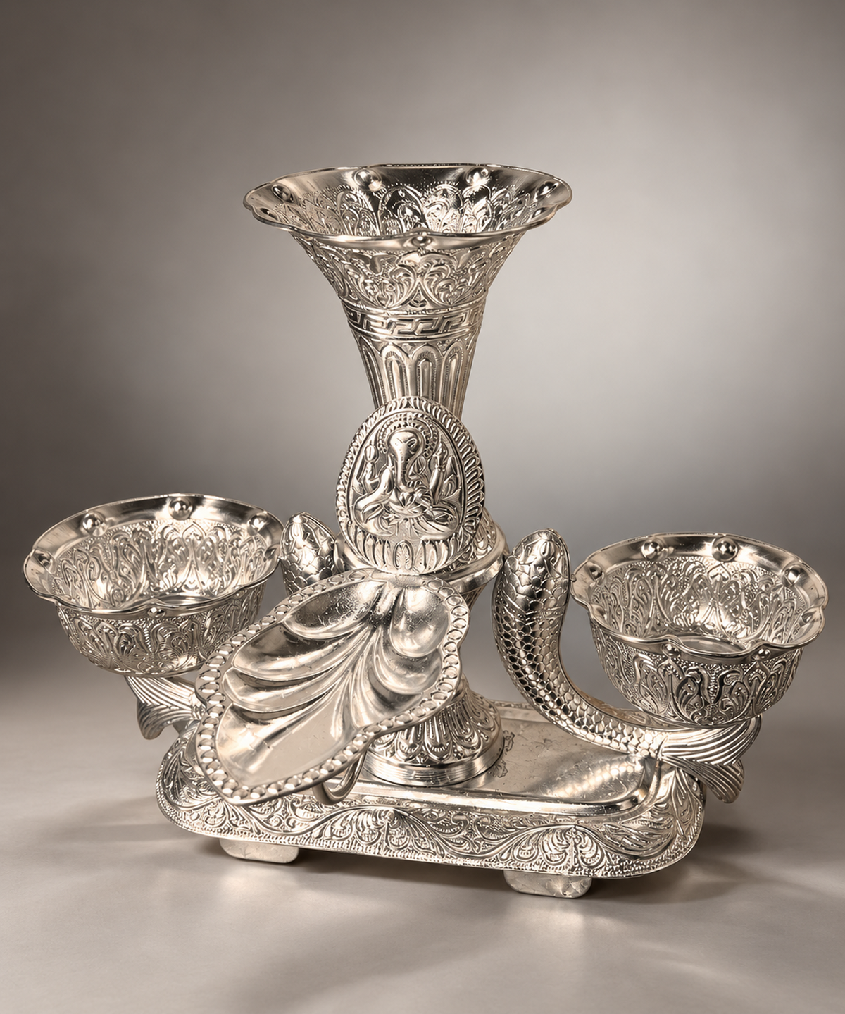An ornate silver-toned metal candelabra with a central flared stem, three bowl-shaped candle holders, and detailed relief carvings, photographed on a neutral gray studio background.
