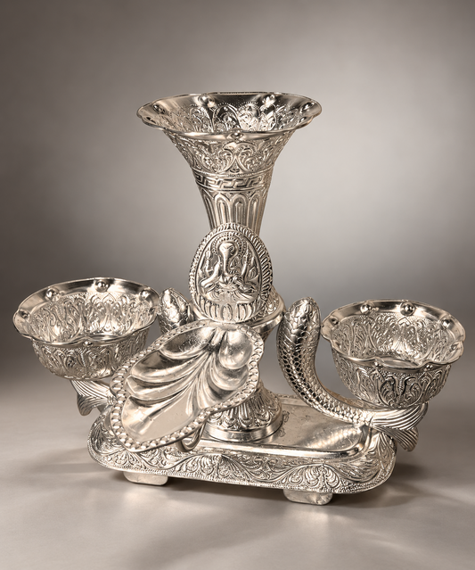 An ornate silver-toned metal candelabra with a central flared stem, three bowl-shaped candle holders, and detailed relief carvings, photographed on a neutral gray studio background.